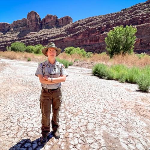 Liz Ballenger stands on a dry floodplain at the southern edge of Arches National Park, Aug. 19, 2024. A lone cottonwood tree rises from the brush behind her near the Colorado River. David Condos/KUER