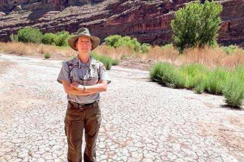 Liz Ballenger stands on a dry floodplain at the southern edge of Arches National Park, Aug. 19, 2024. A lone cottonwood tree rises from the brush behind her near the Colorado River. David Condos/KUER